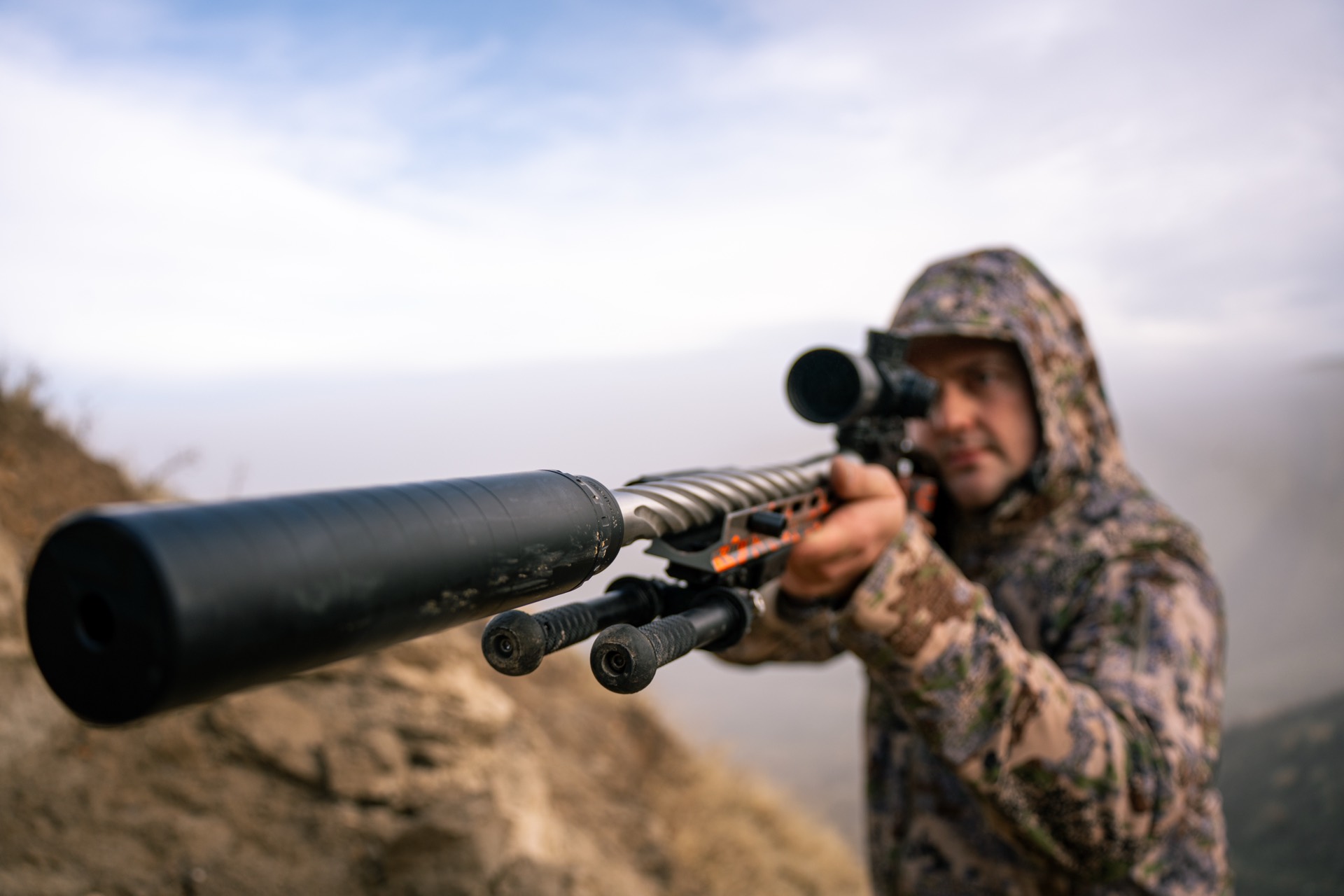 Hunter in camo aiming a suppressed rifle from a ridgeline